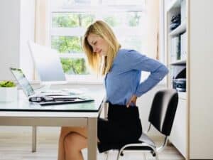 An image of a person sitting at a desk for prolonged hours, visibly fatigued and holding their lower back in discomfort, highlighting the impact of prolonged sitting on lower back health.